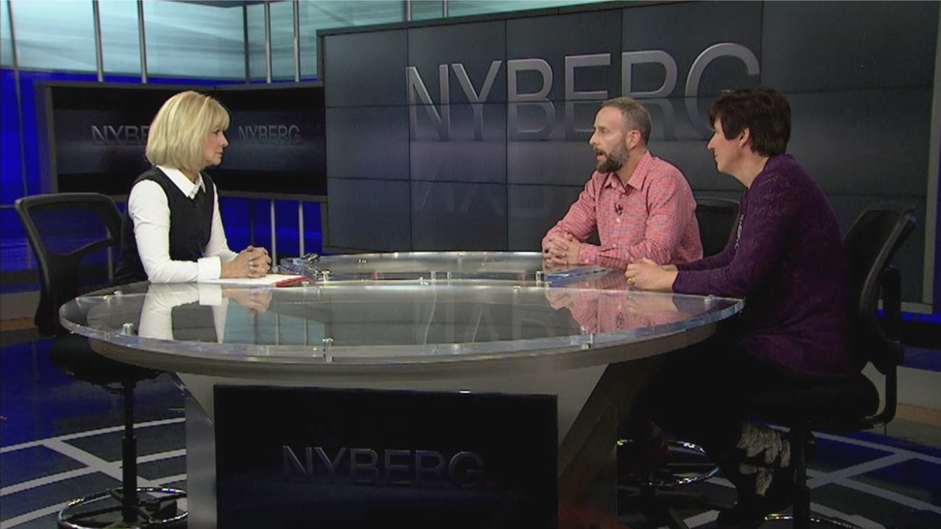 Three people sitting at a round glass table in a TV studio for a discussion or interview, with the NYBERG logo visible on screens in the background.