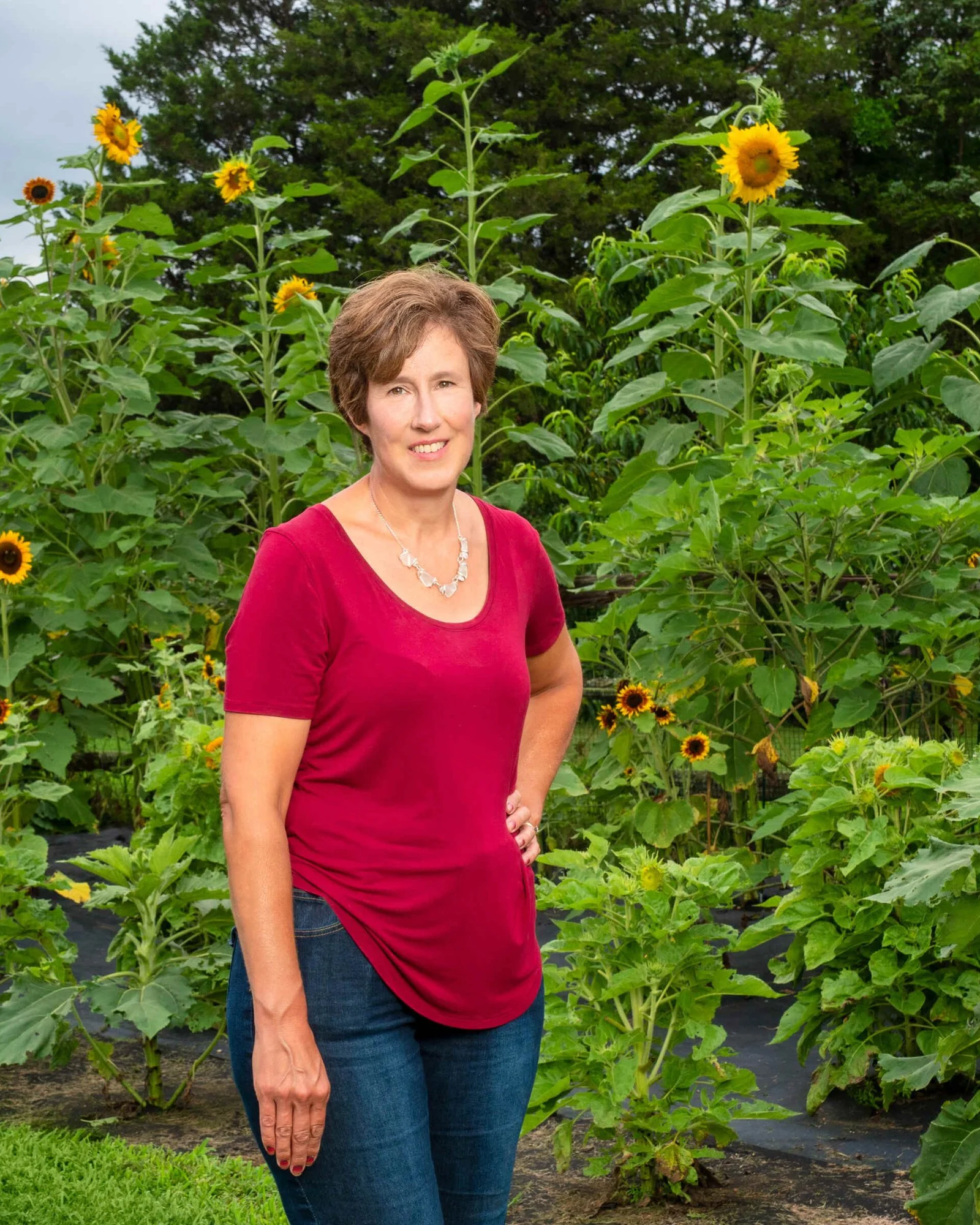 A woman standing in a sunflower field with greenery in the background, wearing a red shirt and blue jeans.