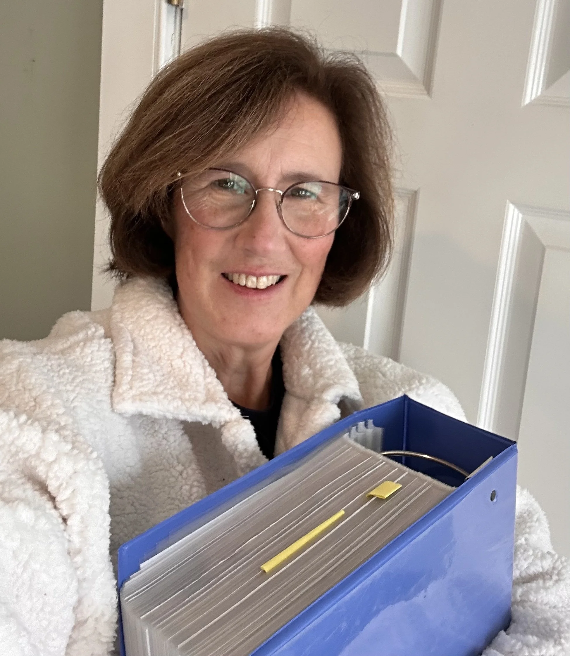 A middle-aged woman with glasses and short brown hair smiling and holding a blue binder filled with papers.