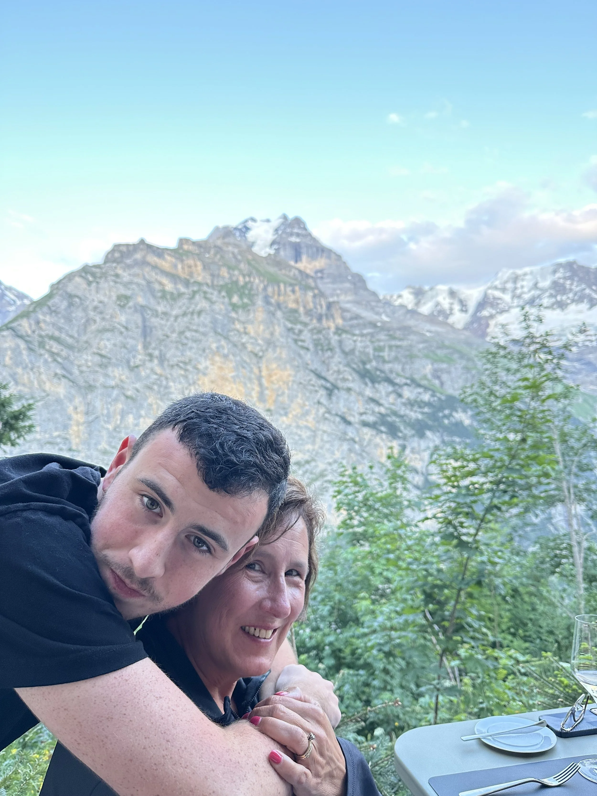 A young man and an older woman hugging at an outdoor table with a mountain landscape in the background.