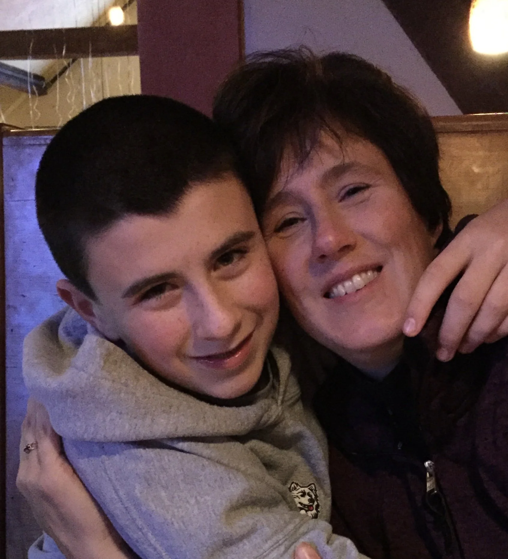 A boy and his mom hugging and smiling in a cozy, dimly lit room.