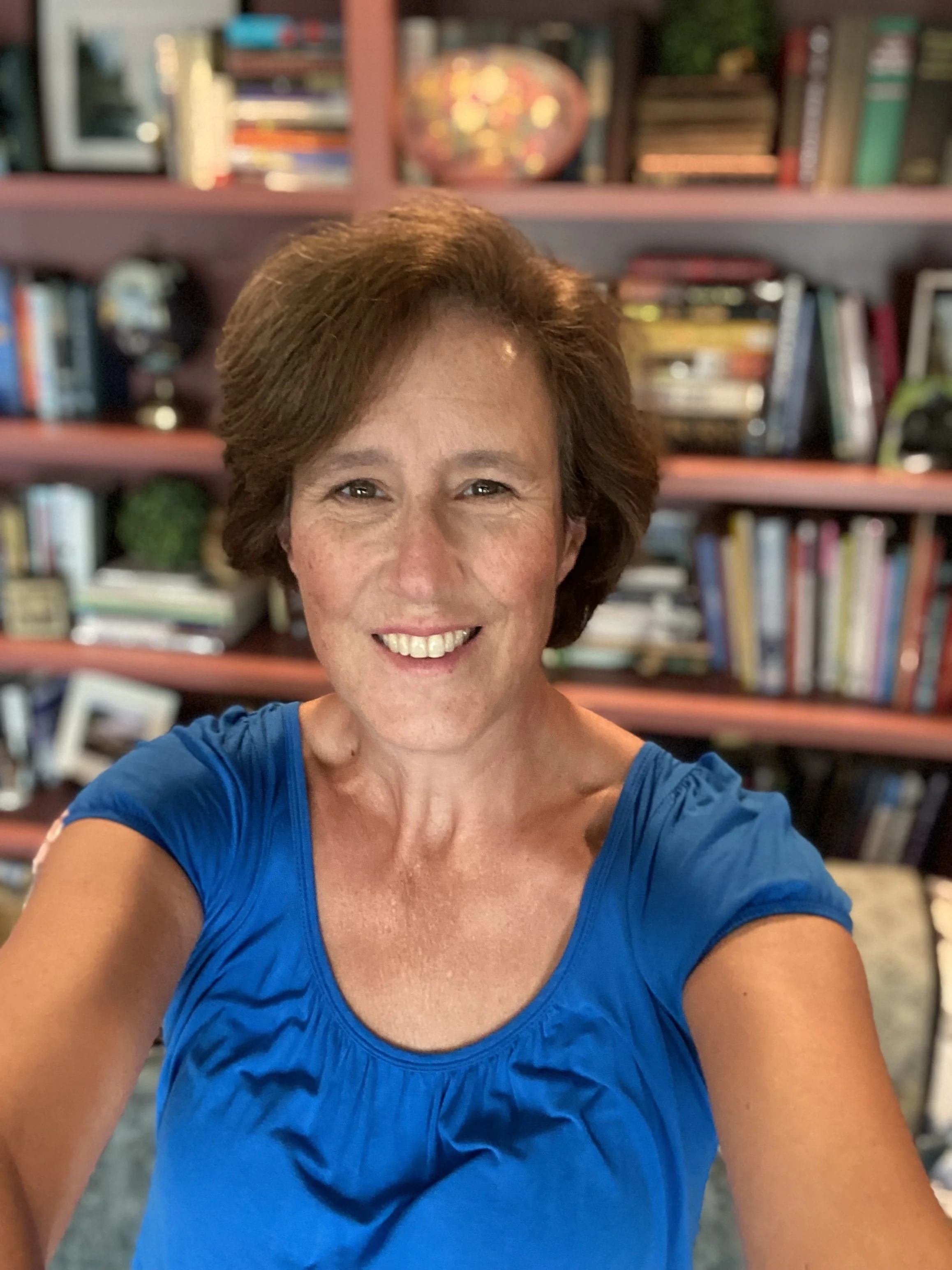 A smiling woman with short brown hair wearing a blue top, taking a selfie in front of a bookshelf filled with books and decorative items.