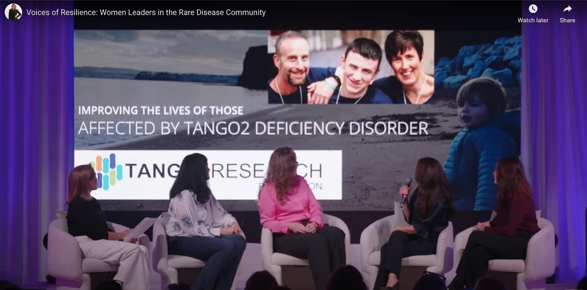 Panel discussion on stage with five women seated in white chairs, one of whom is speaking into a microphone. Behind them is a large screen displaying an image of a family and text about research on Tang2 deficiency disorder.