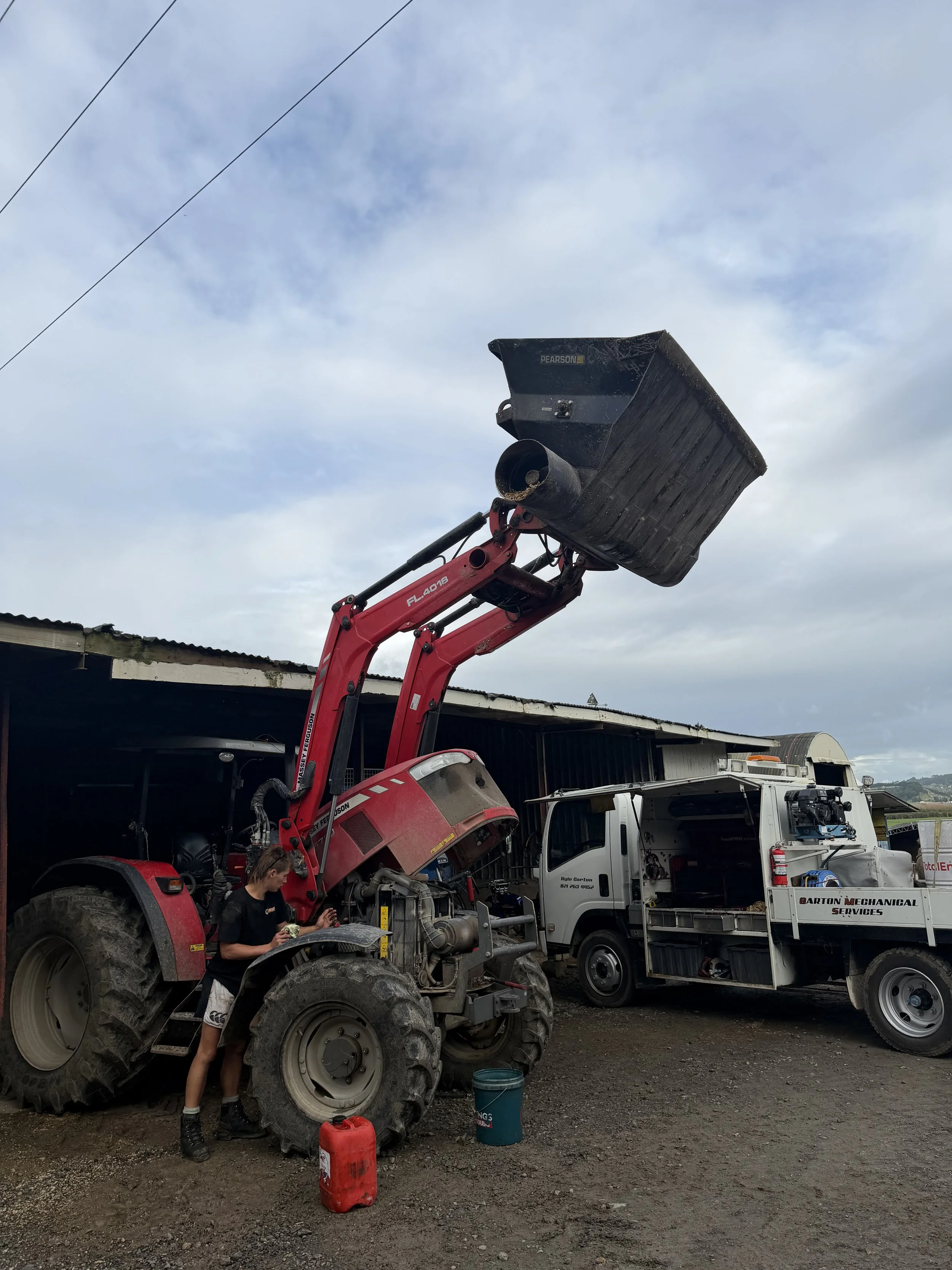 A diesel mechanic working on a tractor at a service station with a mechanic truck nearby. The mechanic is holding tools and is near the tractor's engine. The service area has a black shed and a cloudy sky.