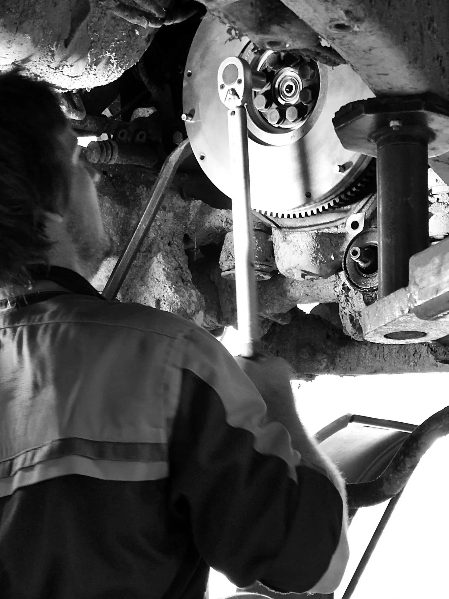 A person working on car engine or transmission, using a wrench, in a garage or workshop, in black and white.
