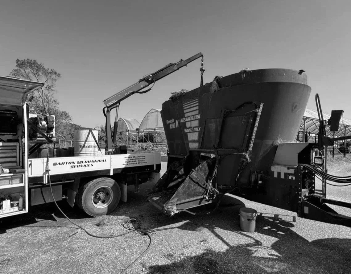 A large boat being lifted by a tow truck with a crane arm attached to the boat's front. The boat is on land, with trees and structures in the background, and is black and white.