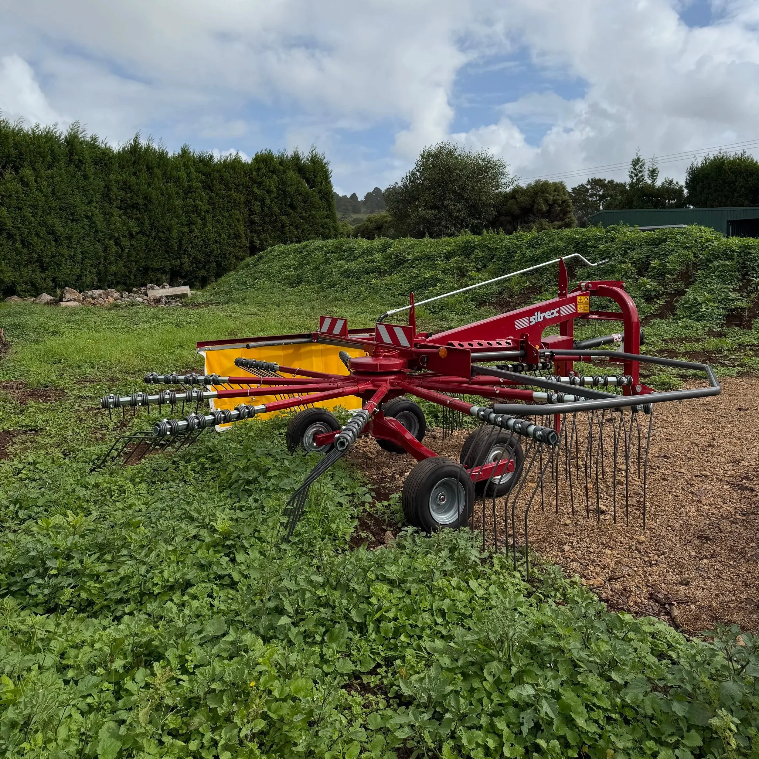 A red and yellow farm rake machine working in a green field with trees and a partly cloudy sky in the background.