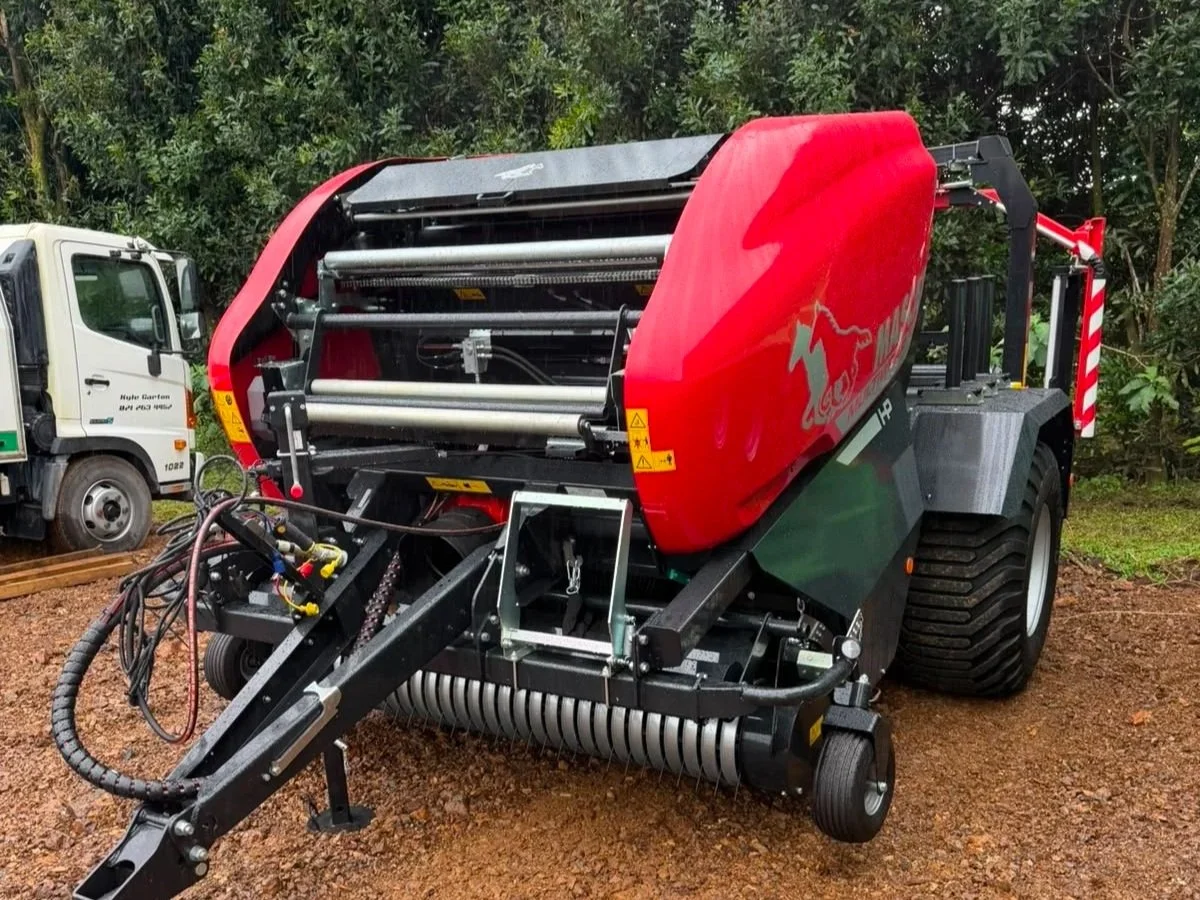 A large industrial hay baler machine with a red body and black frame outdoors on dirt ground, surrounded by green trees.