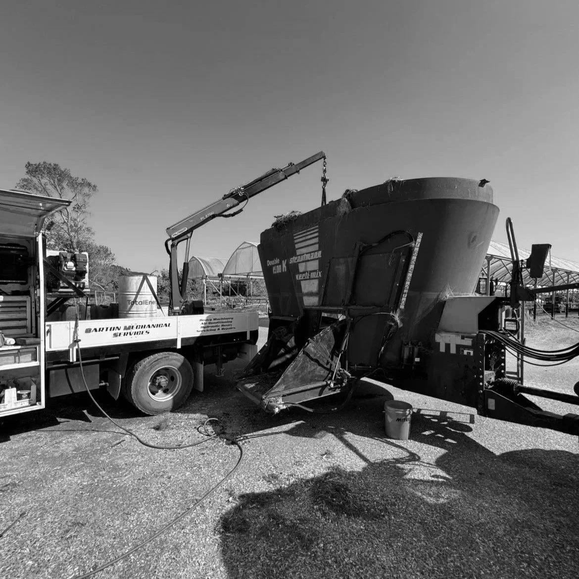 A large industrial machine with a hopper attached, connected to a truck labeled 'Barton Mechanical Services,' in an outdoor agricultural setting with greenhouses in the background.