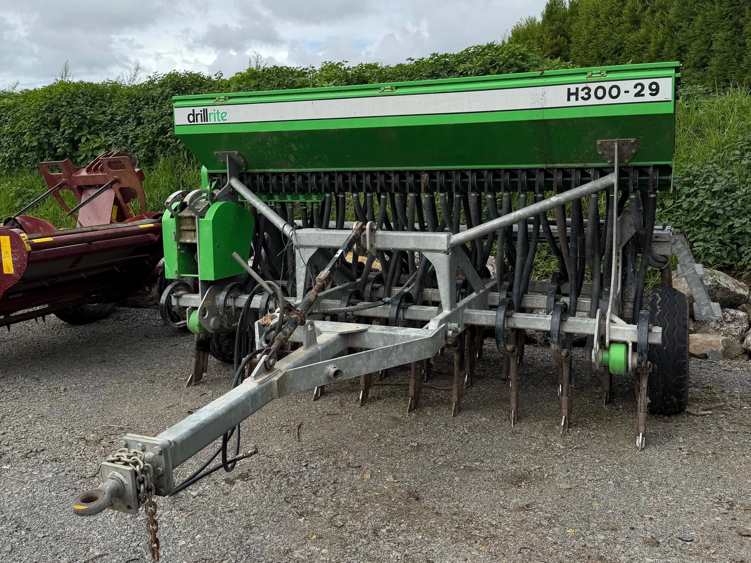 Green agricultural seed drill machine with black hoses and metal parts parked outdoors on gravel with green bushes and cloudy sky in the background.