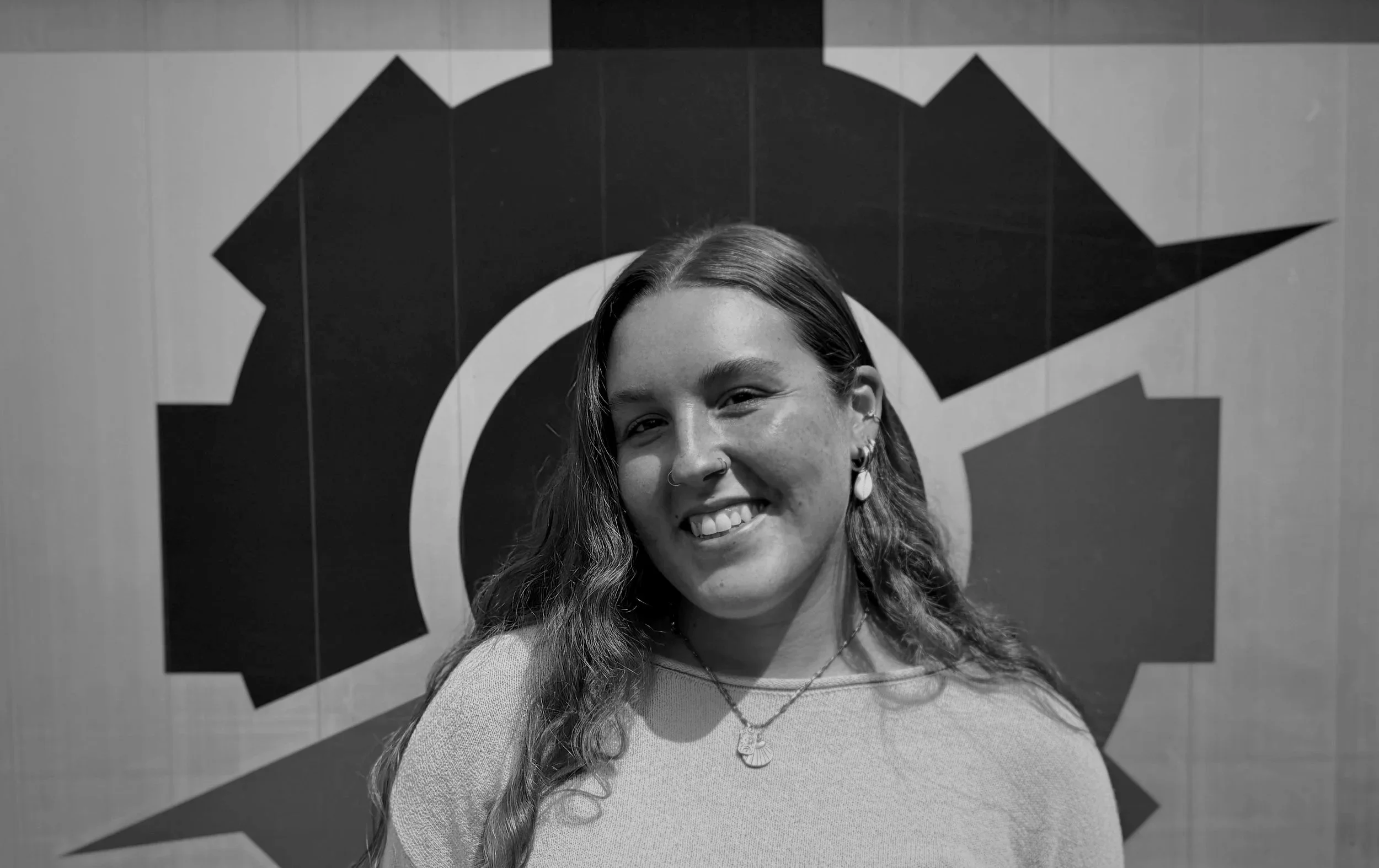 Portrait of a smiling young woman with long wavy hair, wearing earrings, necklaces, and a light-colored top, standing in front of a wall with a large abstract geometric design.