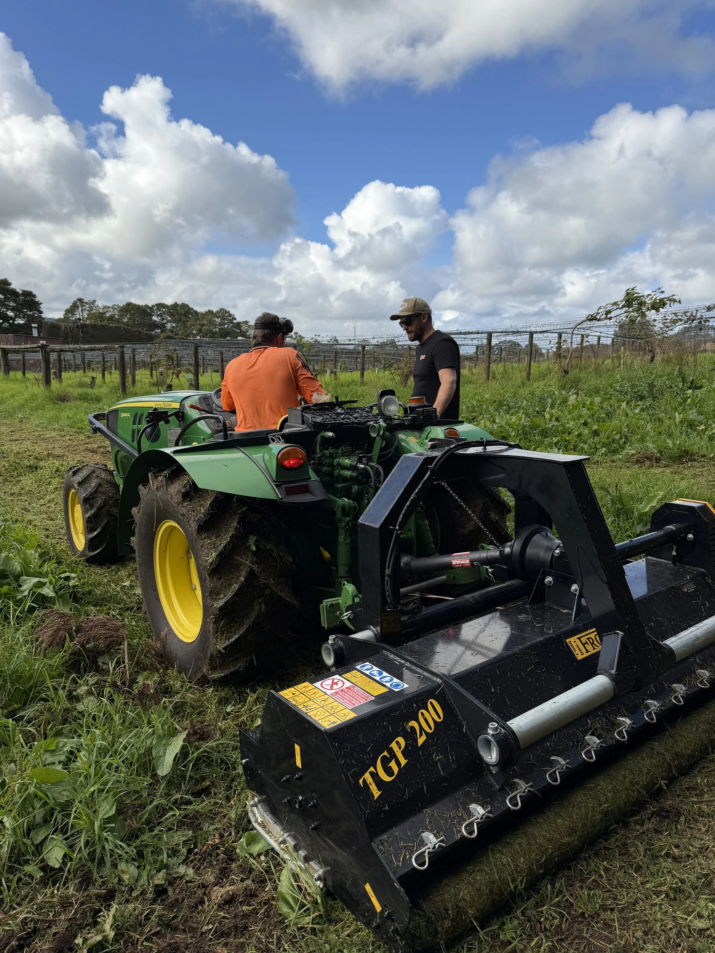 Two mechanics are working with a green John Deere tractor outdoors in a field with green grass and plants, with a partly cloudy sky overhead.