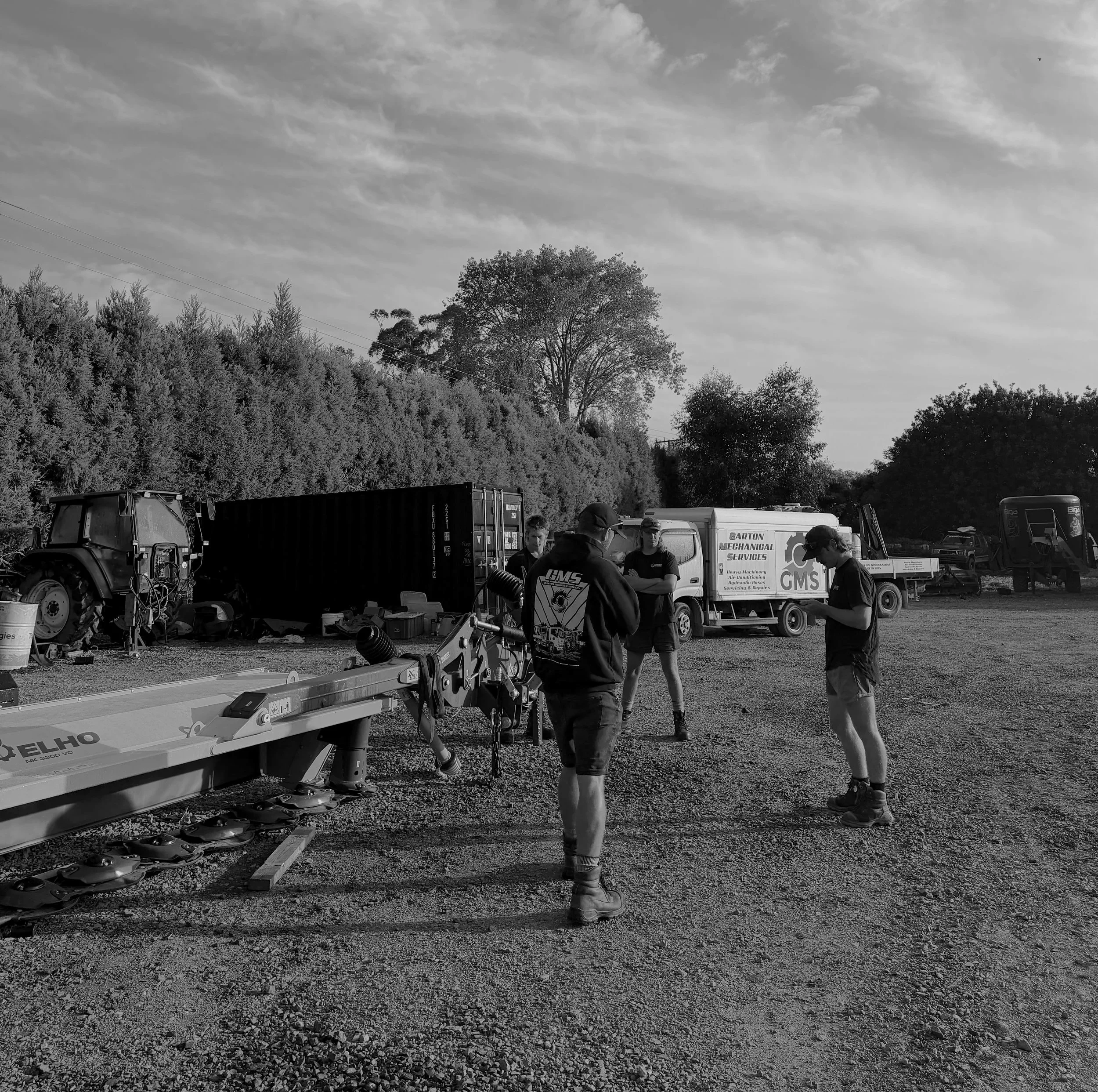 Three people standing outdoors near construction and machinery equipment, with trucks, containers, and trees in the background, under a partly cloudy sky.