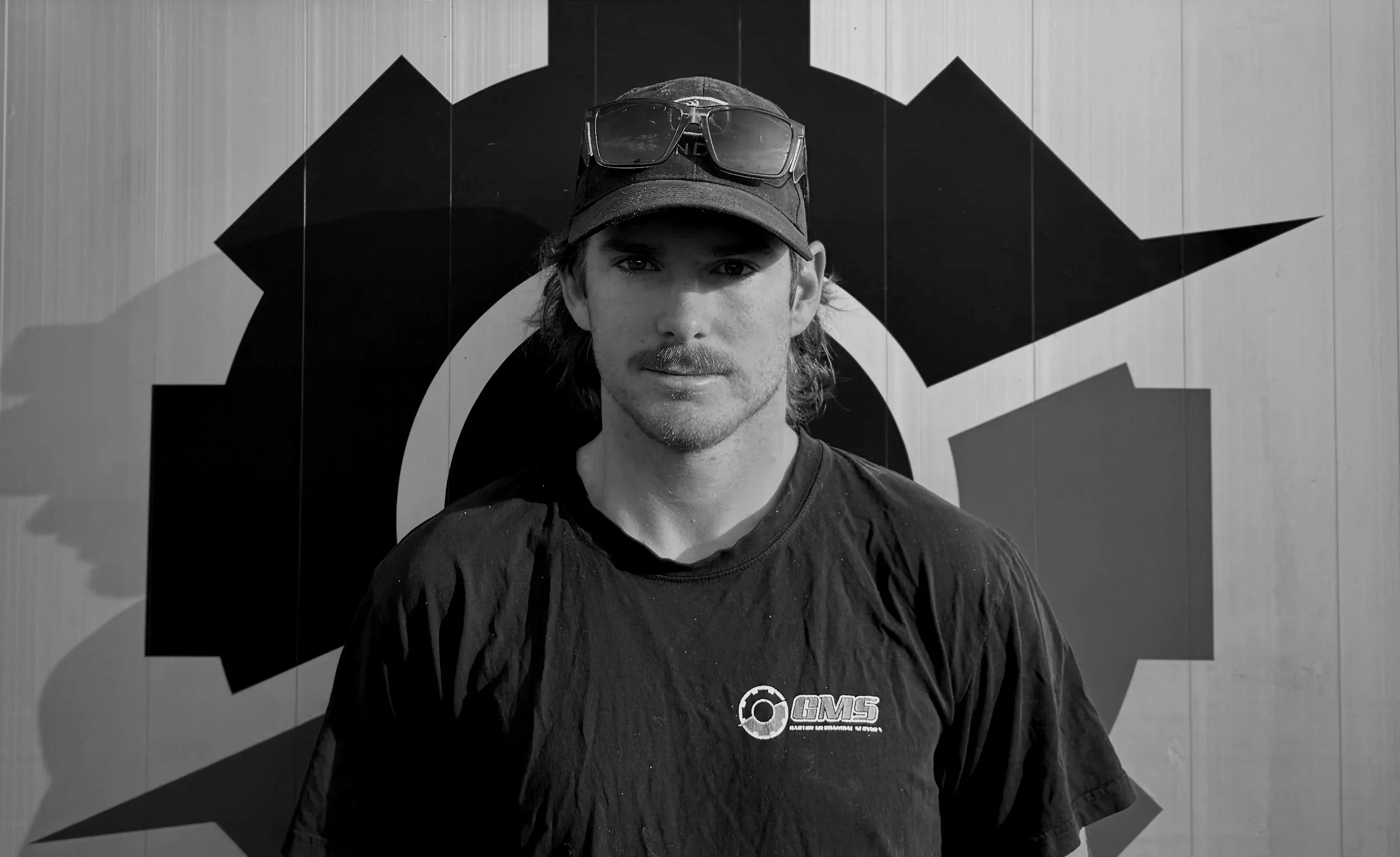 Young man wearing a cap and sunglasses on his head, standing outdoors in front of a mural with geometric shapes.