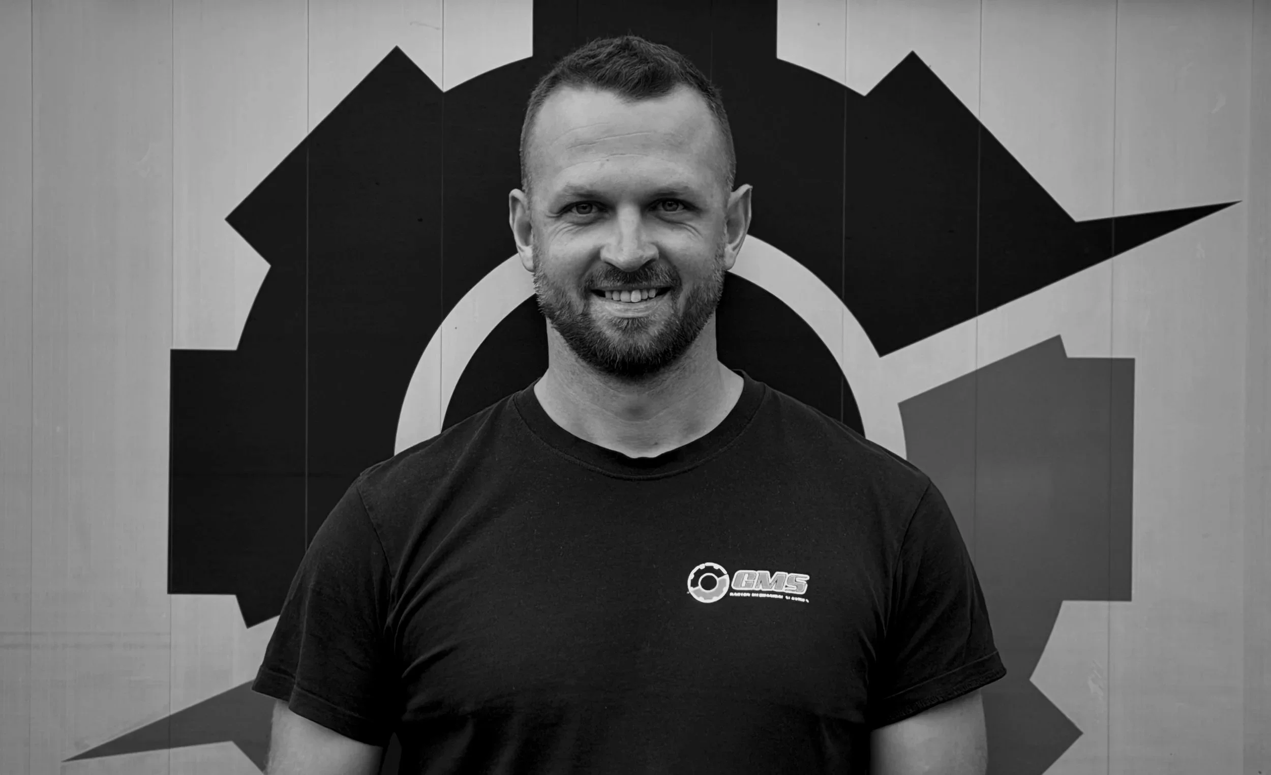 A smiling man with a beard and short hair wearing a black t-shirt with a logo, standing in front of a geometric background with a gear and arrow design.
