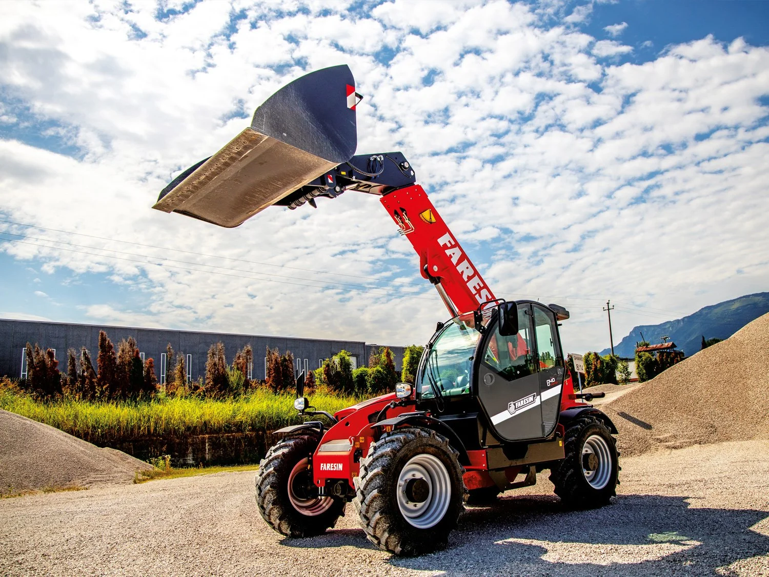 A red and black telescopic forklift with a large scoop attachment, branded 'FARESIN,' parked outdoors on gravel with a backdrop of mountains, trees, and a partly cloudy sky.