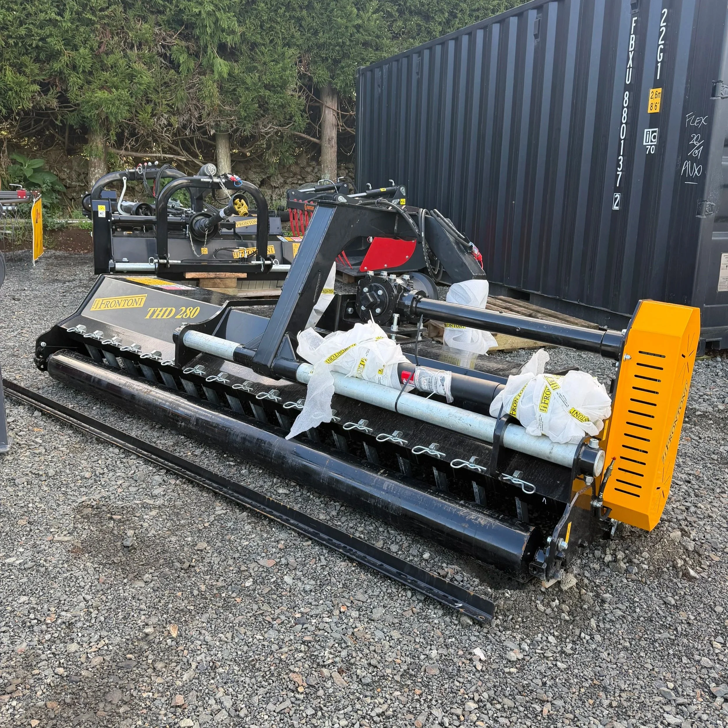 Construction equipment and a large shipping container placed on gravel with trees in the background.
