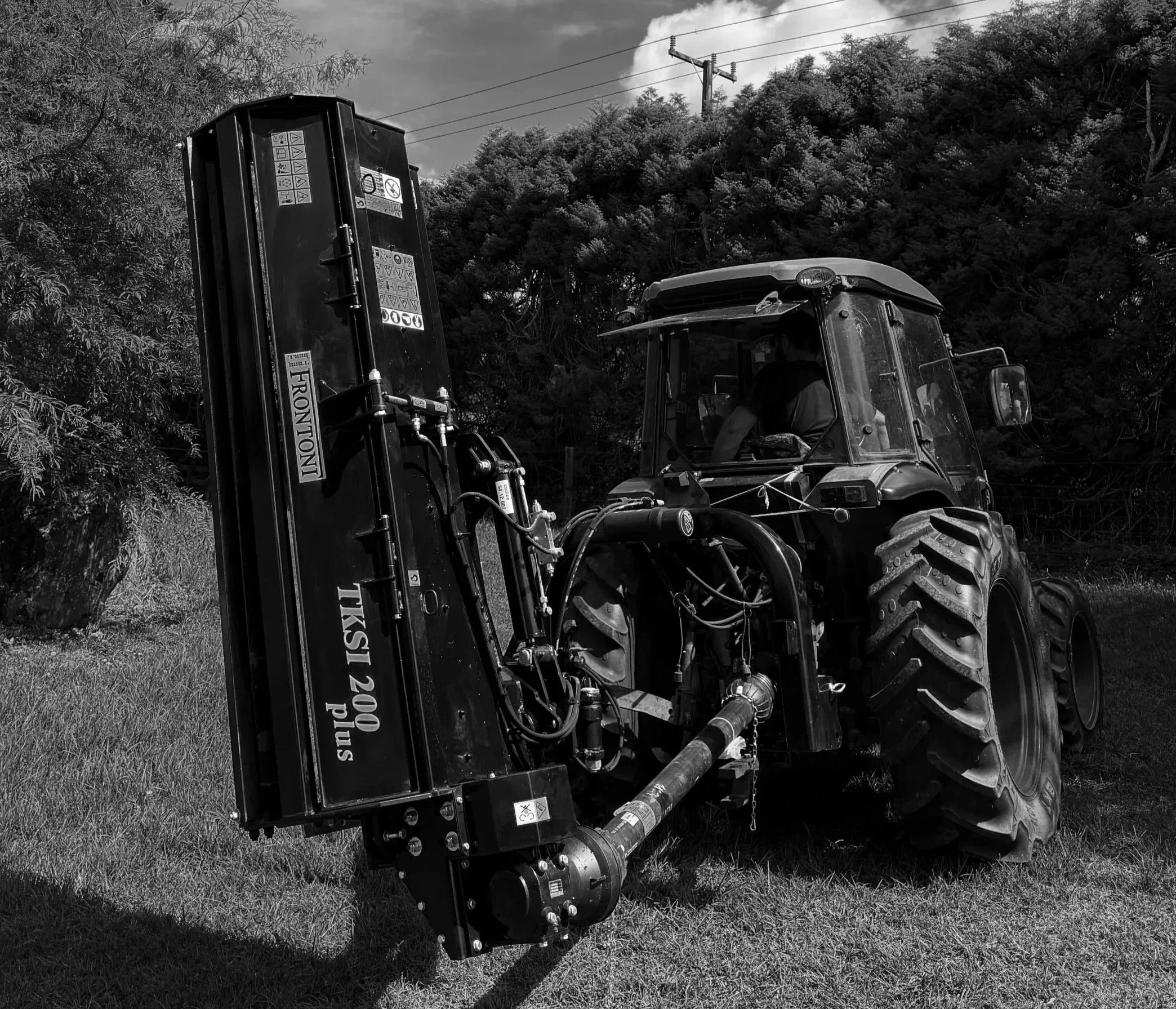 A tractor equipped with a large mower attachment on a grassy area, with trees and power lines in the background.