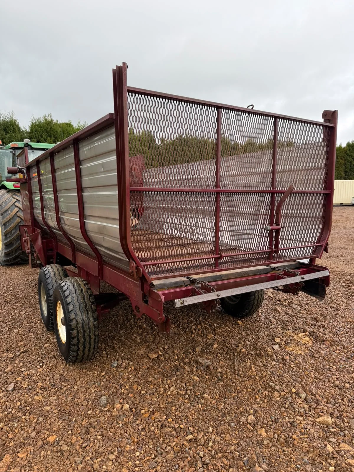 Red farm grain trailer with a metal mesh gate and large tires on gravel ground, overcast sky in the background.