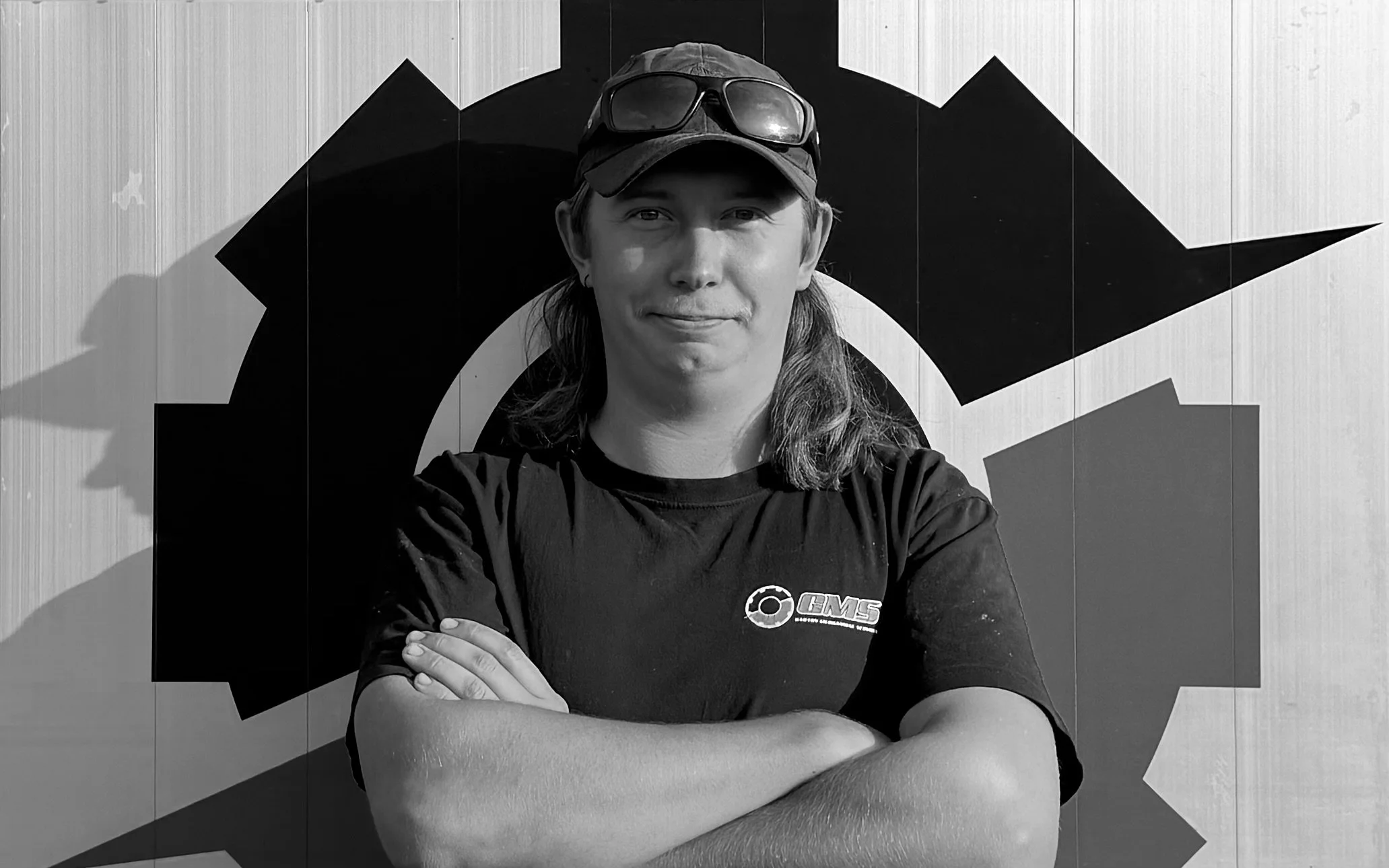 A young man with long hair, wearing a cap and sunglasses on his head, stands with arms crossed in front of a graphic mural on a wall. 