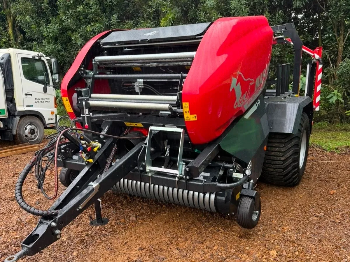 A large red and black specialized agricultural or industrial equipment on a dirt surface, with a white truck in the background and green trees behind.