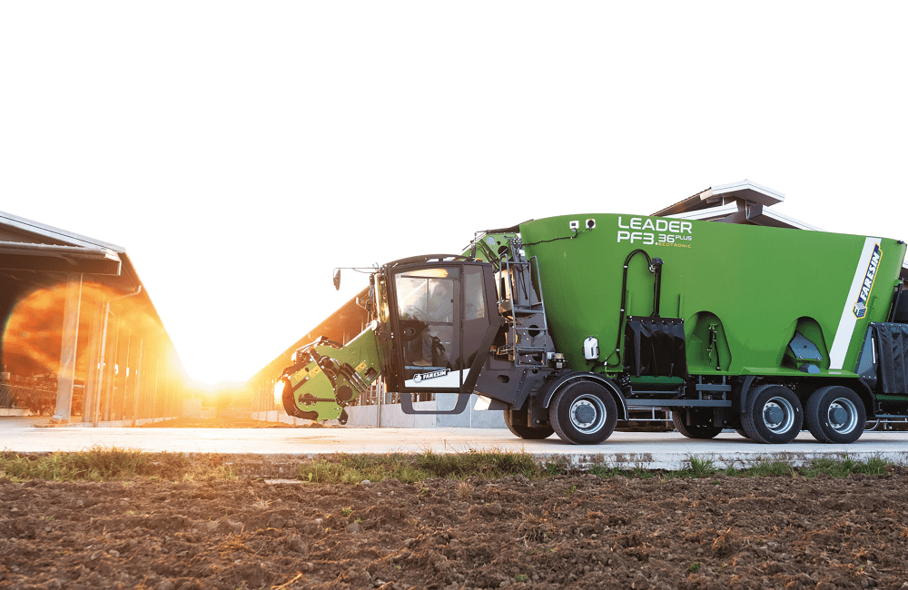 A green street sweeper vehicle operating outdoors at sunset, with a building on the left side.