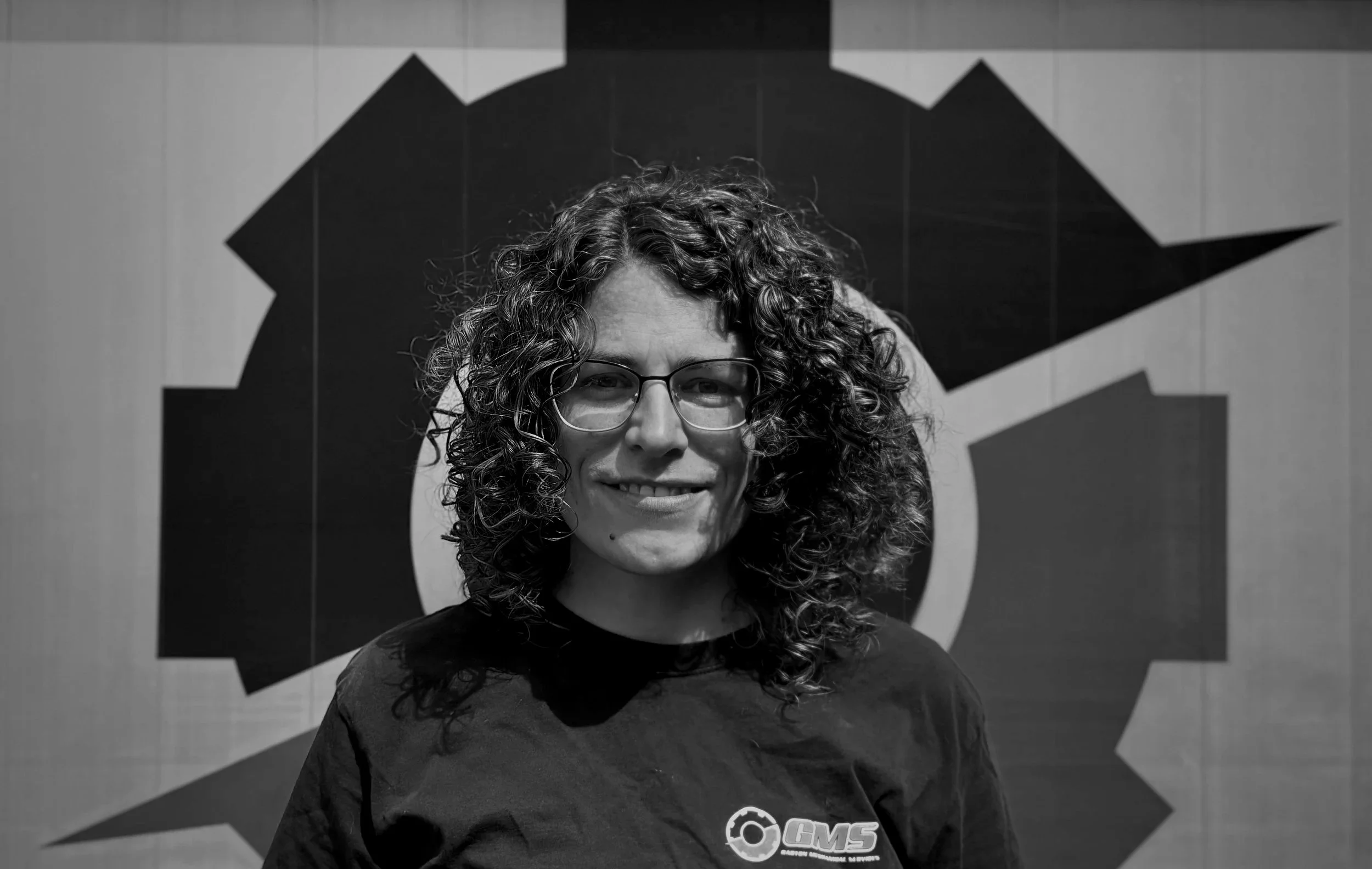 Black and white photo of a woman with curly hair and glasses, smiling, standing in front of a gear-shaped symbol on a wall.