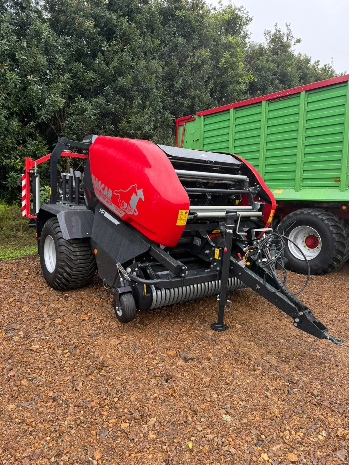 A large agricultural machine with a red cover and black framework, parked on a gravel ground next to a green fence. The machine has large tires and various mechanical parts.
