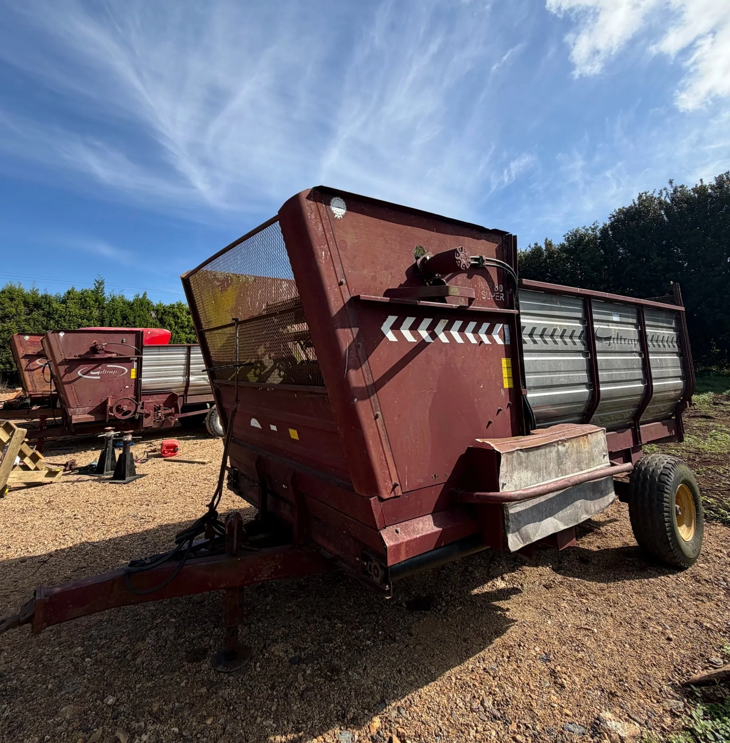 Red wood chipper on a dirt lot with trees in the background under a blue sky with wispy clouds.