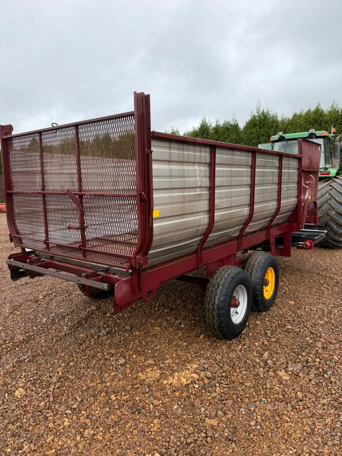 Red farm wagon with metal sides and large tires, attached to a tractor on gravel ground with trees and cloudy sky in the background.