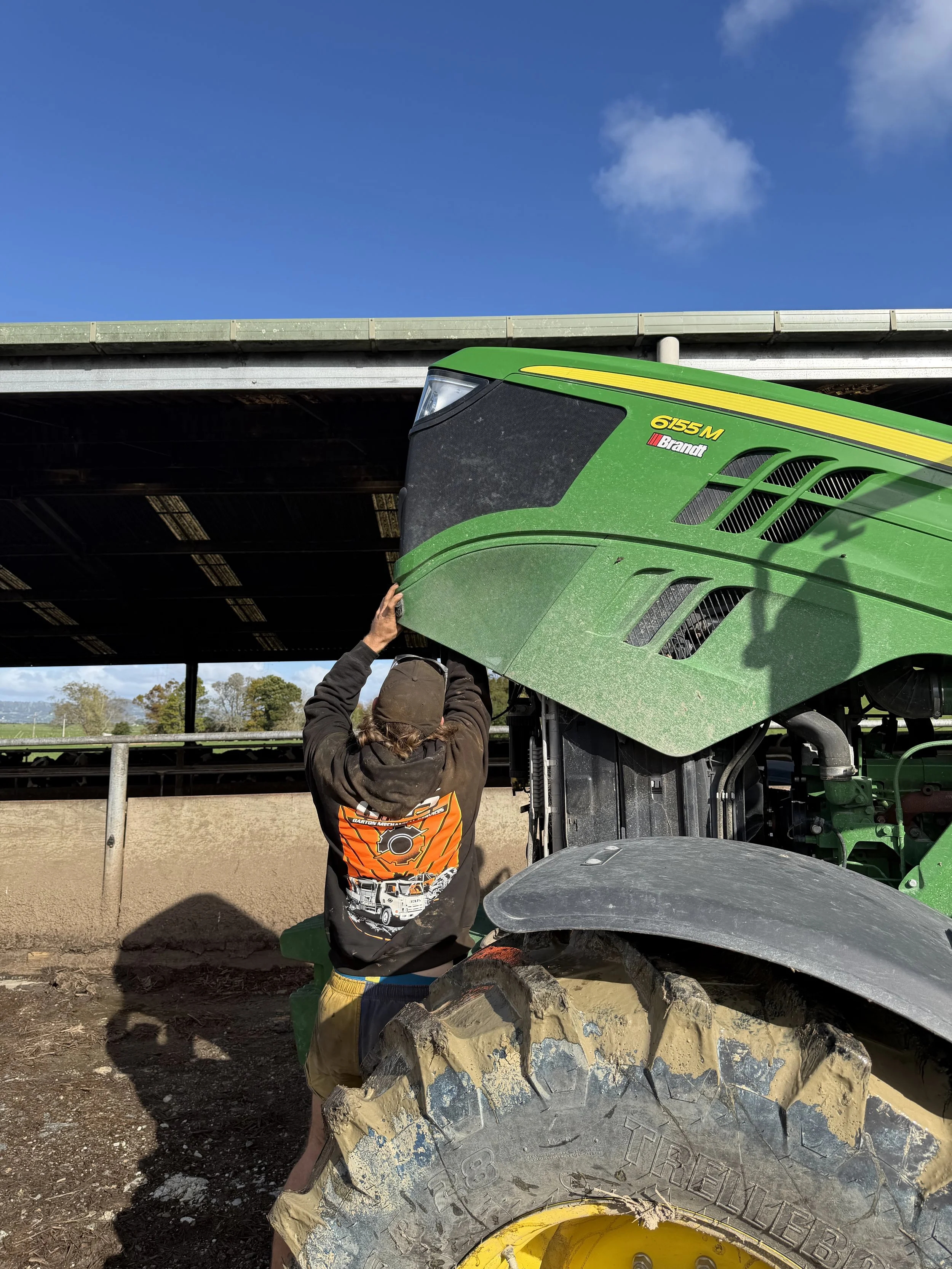 A person wearing a black hoodie with an orange design on the back and yellow pants is standing outdoors, adjusting a large green John Deere agricultural machine under a blue sky with some clouds. There is a large tire in the foreground with mud and dirt on it, and a farm field in the background.