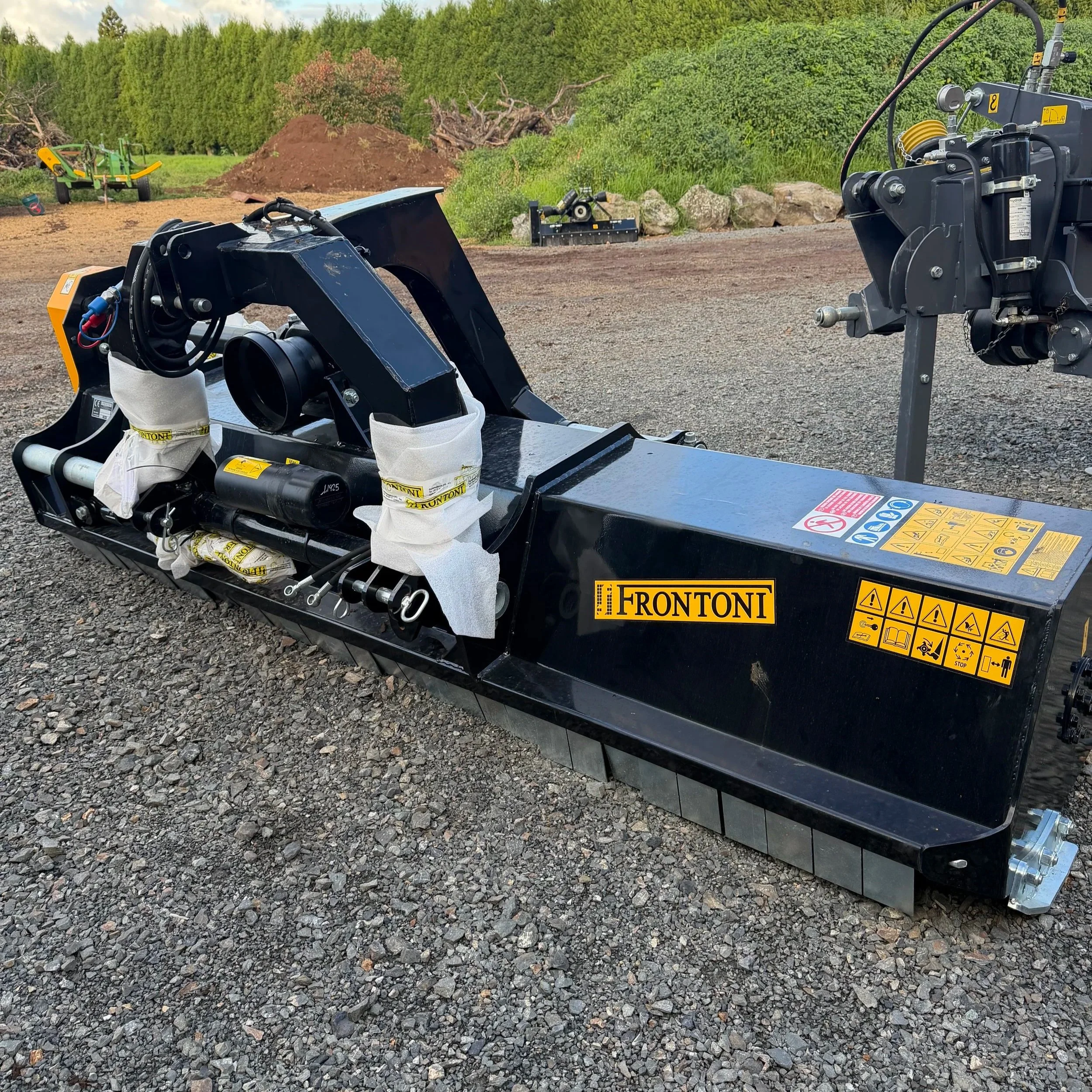 Close-up of construction equipment, including hydraulic machinery with hoses and warning labels, set on gravel ground with green trees and a mound of dirt in the background.