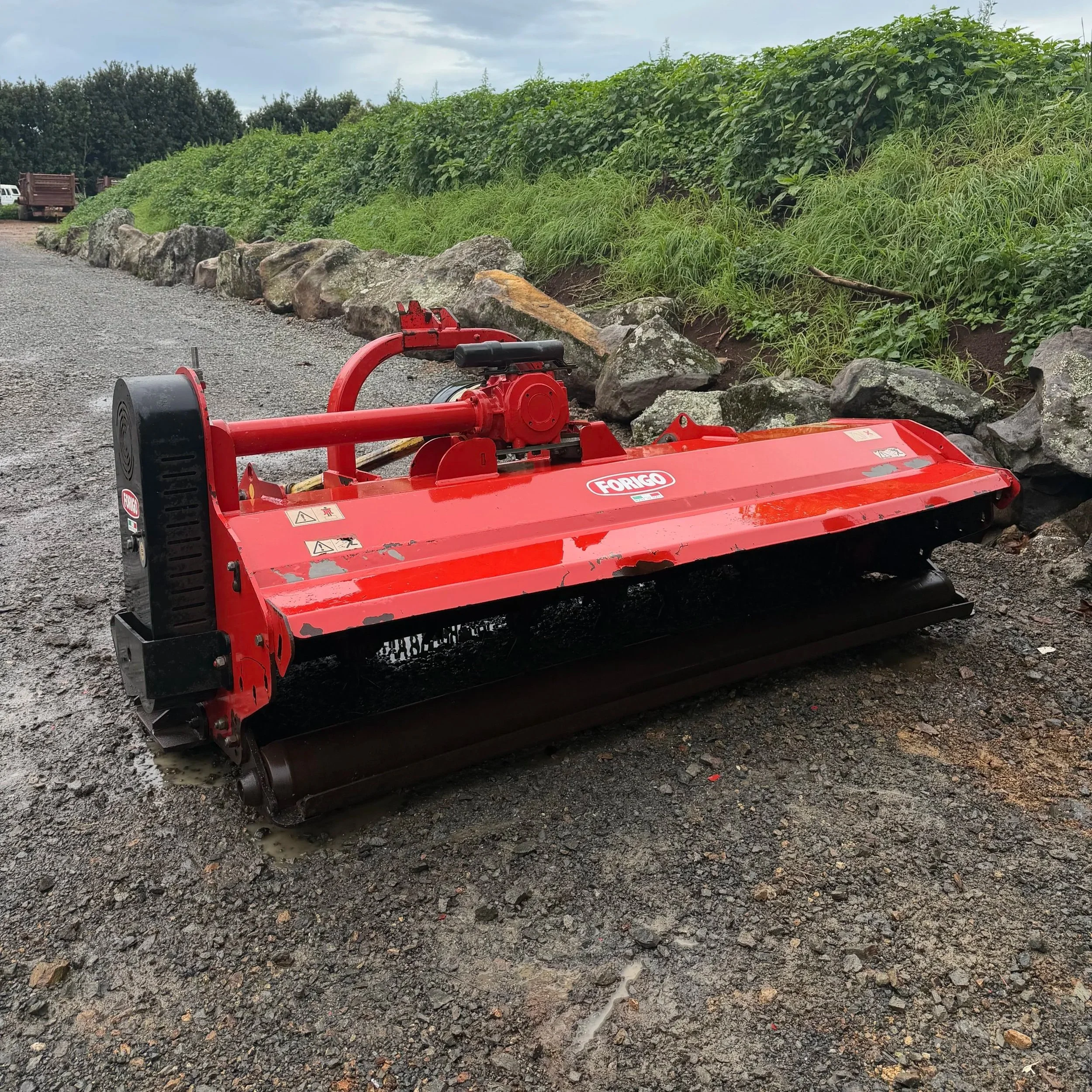 A red Forigo rotary mower attachment for a tractor parked on a gravel surface with rocks and green vegetation in the background.