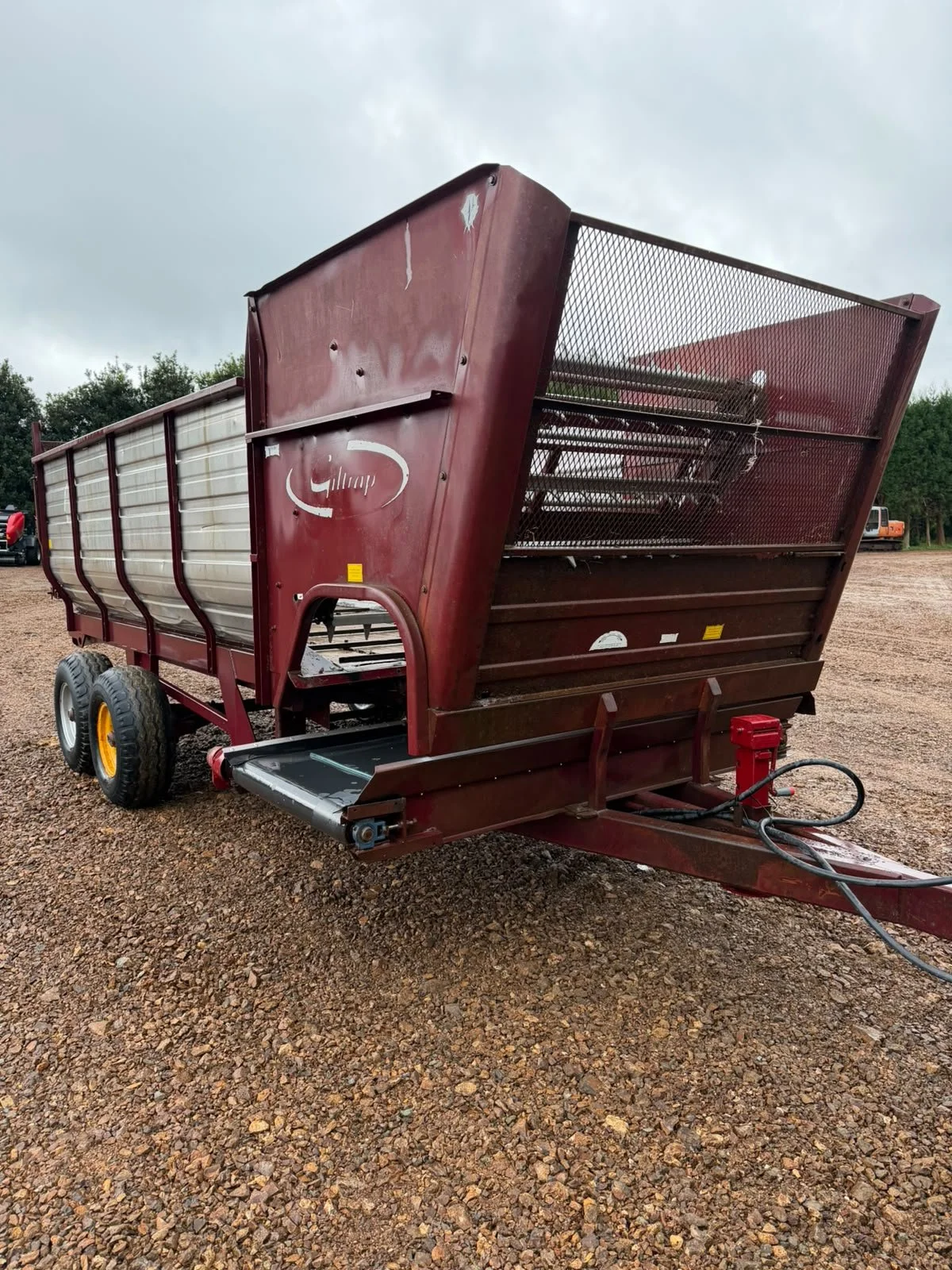 A red livestock trailer is parked on dirt ground, with two wheels visible on the left side, and a hydraulic system at the front. The trailer has a metal mesh section on the right side. The sky is overcast, and trees are visible in the background.