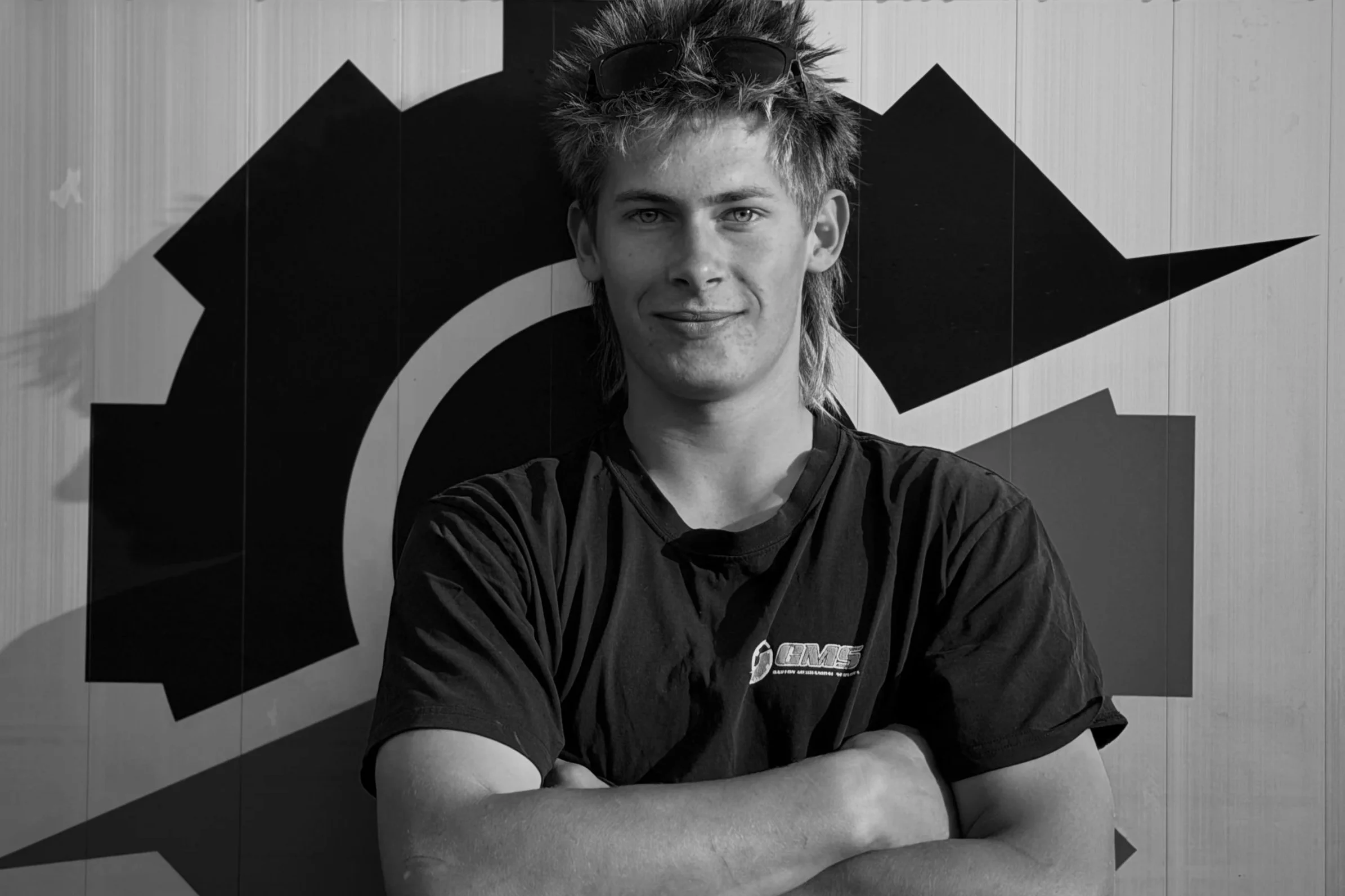 A young man with spiky hair and sunglasses on his head, standing with his arms crossed in front of a wall with a large geometric logo.