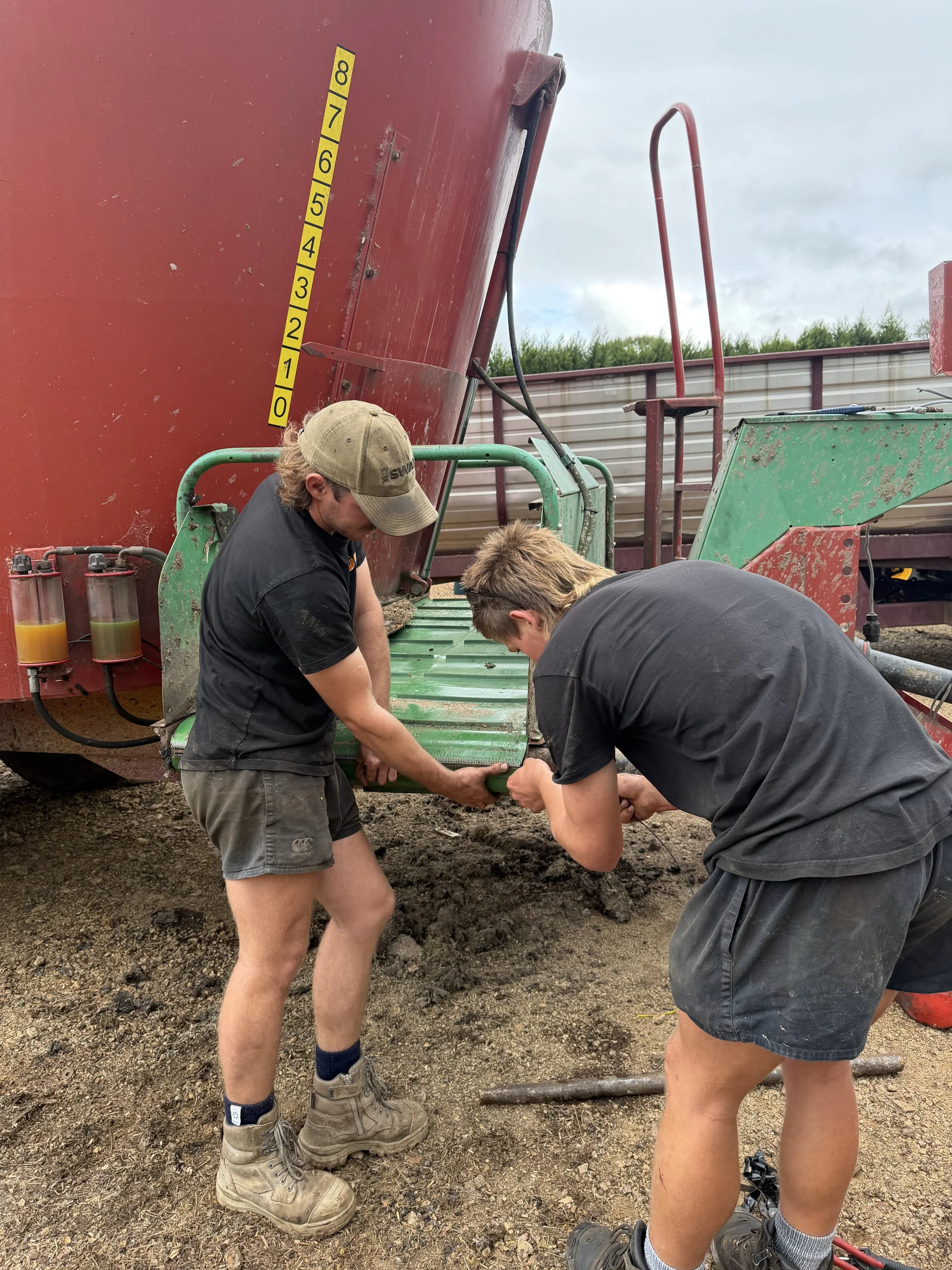 Two diesel mechanics working outdoors next to a large red piece of equipment with a height measurement scale, dirt ground, and a metal fence in the background.