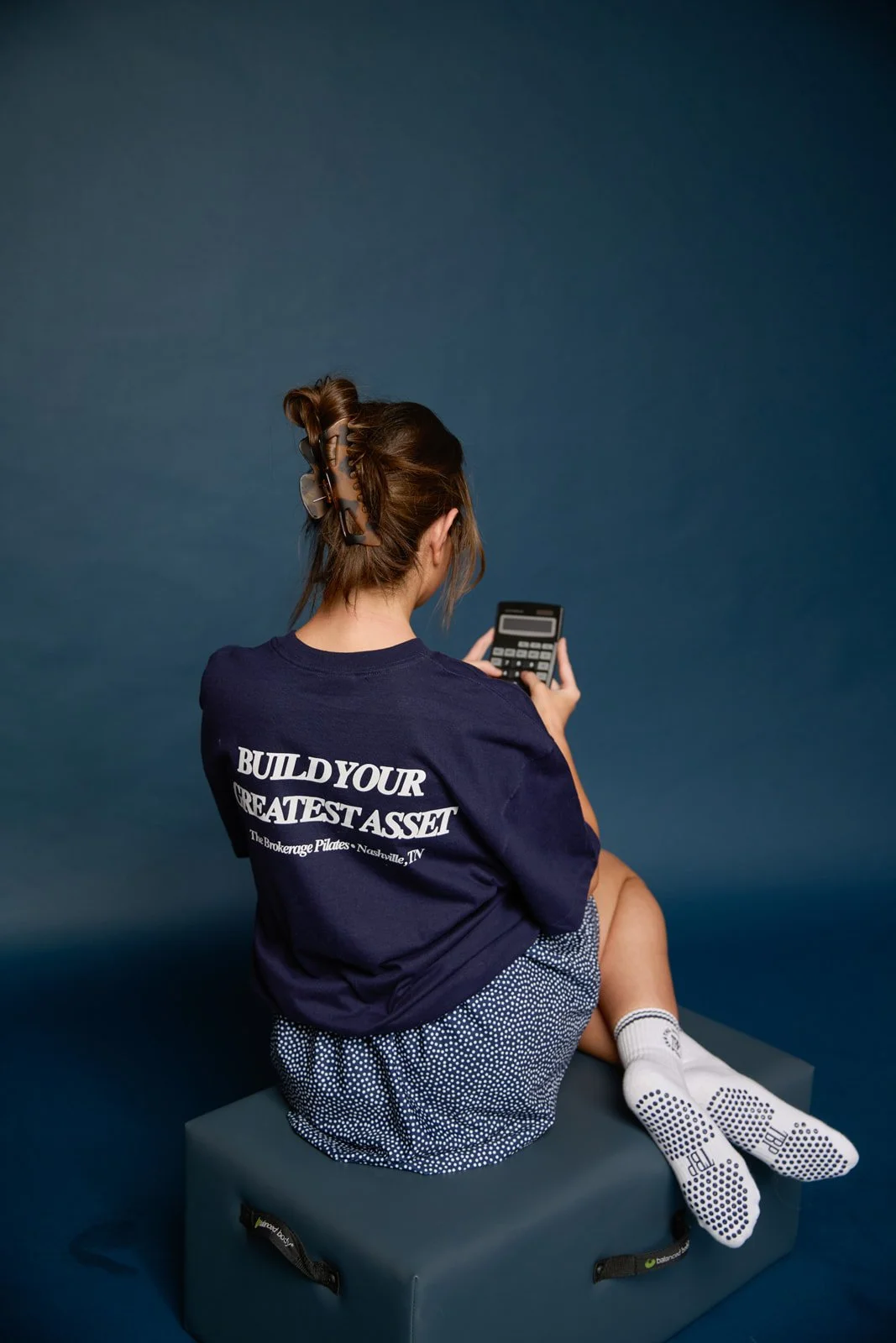 A woman with brown hair in a bun, wearing sunglasses and a navy blue shirt with white text, seated on a gray exercise block, looking at a calculator in her hand, with a plain dark blue background.