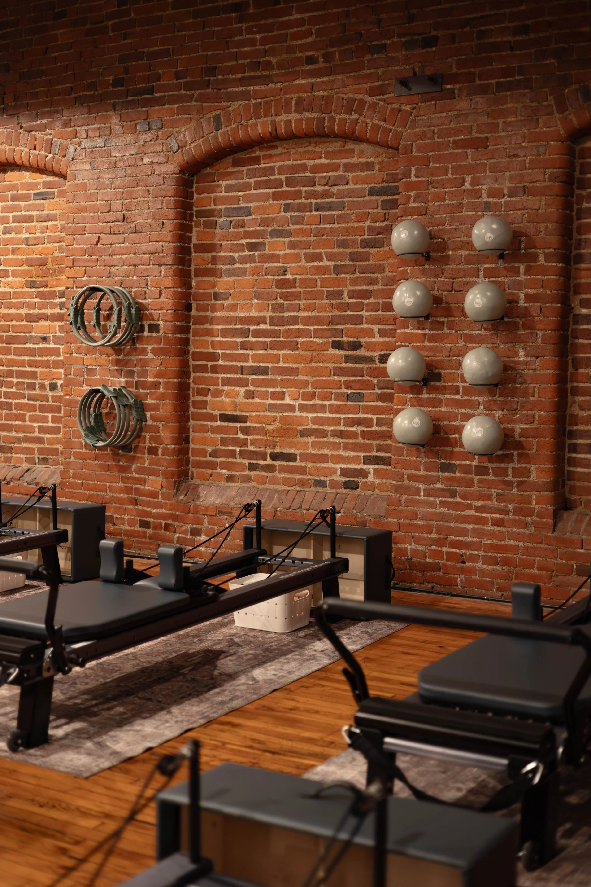 Fitness studio with brick walls, massage rollers on the wall, and Pilates reformers on a rug and hardwood floor.