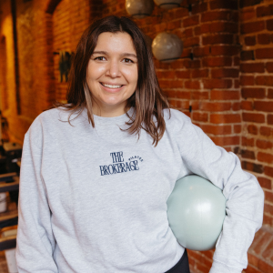 A young woman with shoulder-length brown hair smiling while holding a light blue balloon inside a brick-walled room.