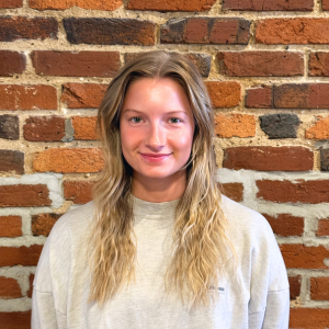 A young woman with long blonde hair wearing a gray sweatshirt, standing in front of a brick wall.