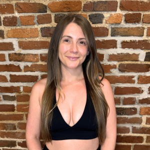 Young woman with long brown hair smiling in front of a brick wall, wearing a black crop top.