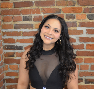A young woman with long, wavy black hair smiling in front of a brick wall.