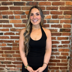A young woman with long, wavy blonde hair wearing a black sleeveless top, standing in front of a brick wall and smiling.