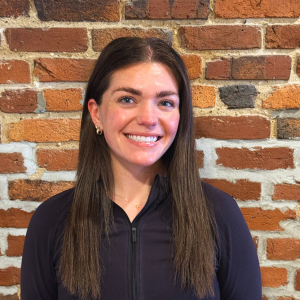 A woman with long brown hair, smiling, wearing a black zip-up jacket, standing in front of a brick wall.