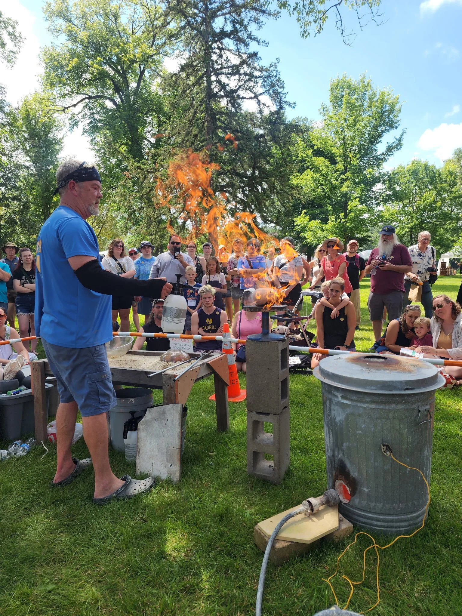 A man performing a science experiment with fire outdoors while a crowd watches, in a park with trees under a clear blue sky.