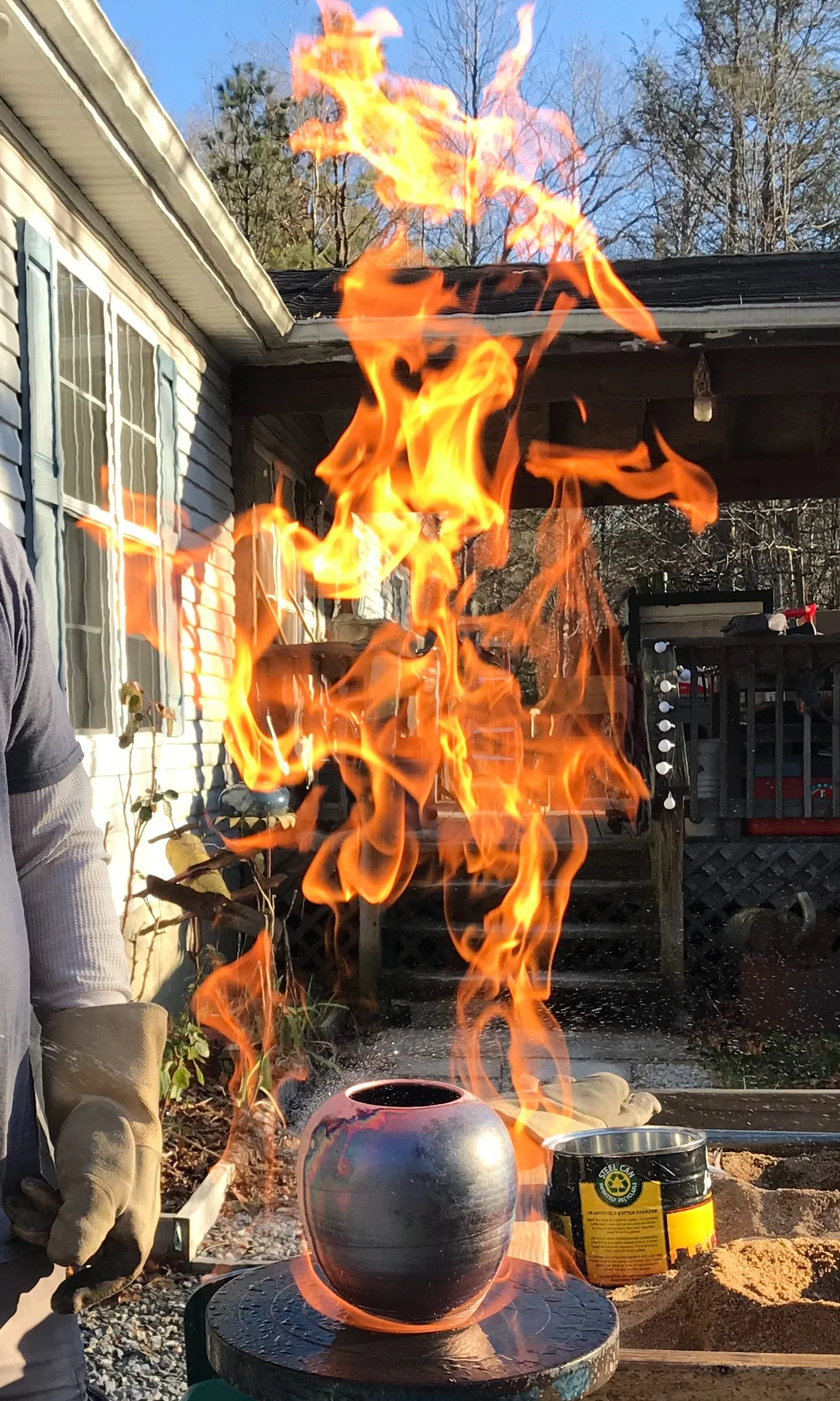 Fire burning inside a ceramic pot on a black table outdoors on a sunny day, with a house, trees, and a deck in the background.