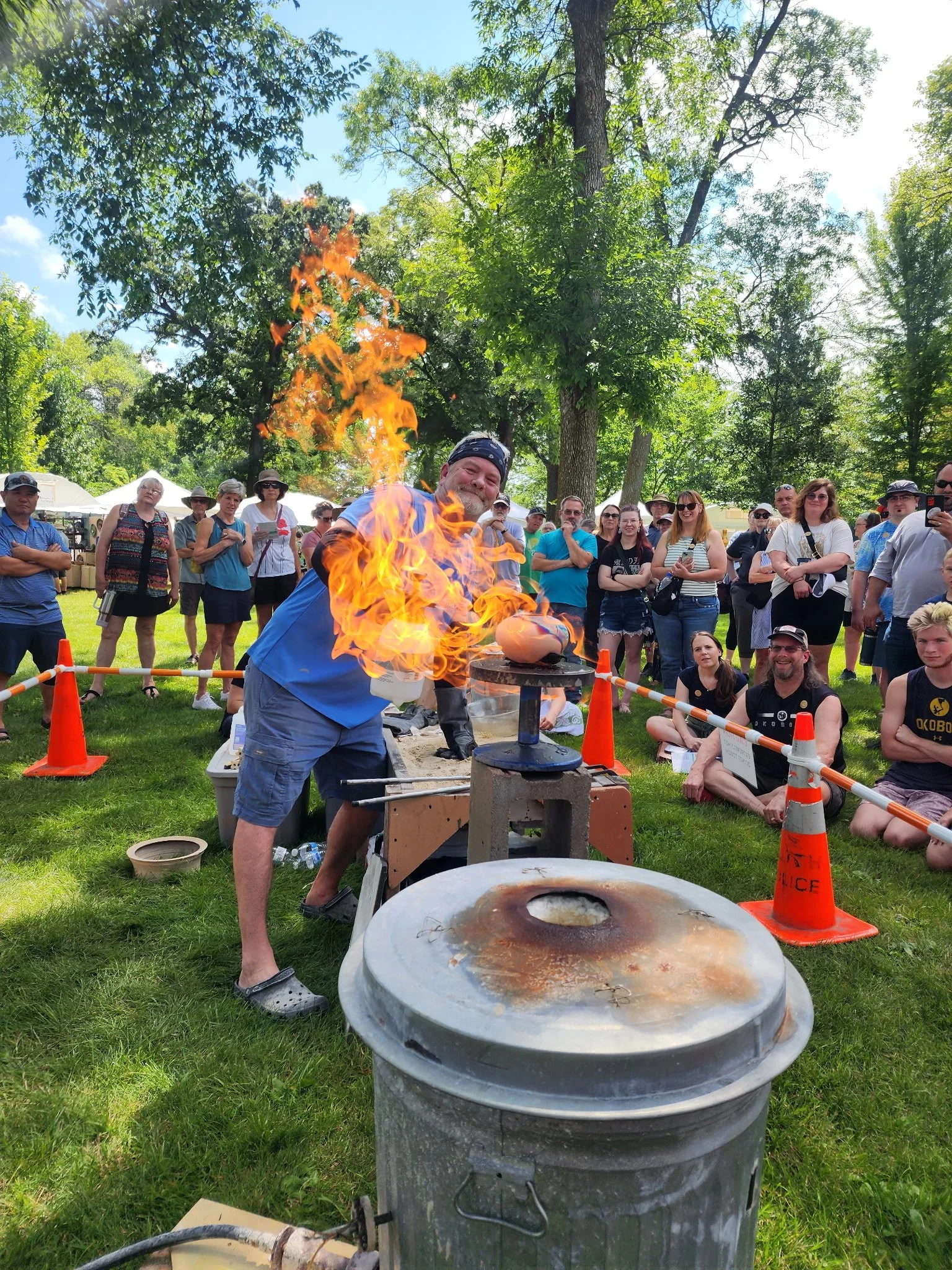 A man performing a science demonstration with fire in a park, surrounded by a crowd of onlookers.