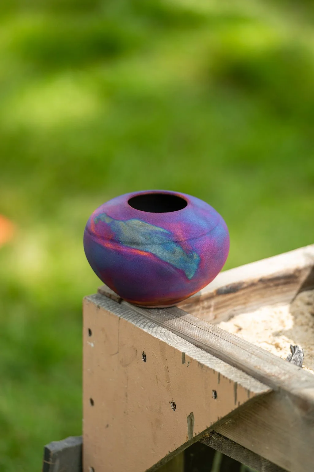 A colorful, round ceramic vase with a narrow opening sits on a wooden surface outdoors, against a blurred green background.
