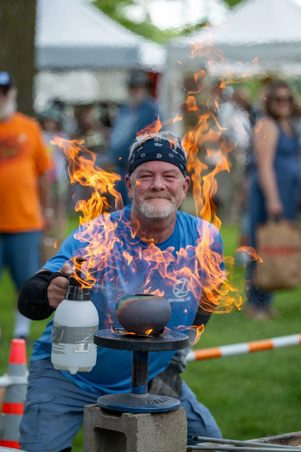 Man with a headband smiling while performing a fire showing stunt with a pot on a metal stand, flames erupting around him at an outdoor event.