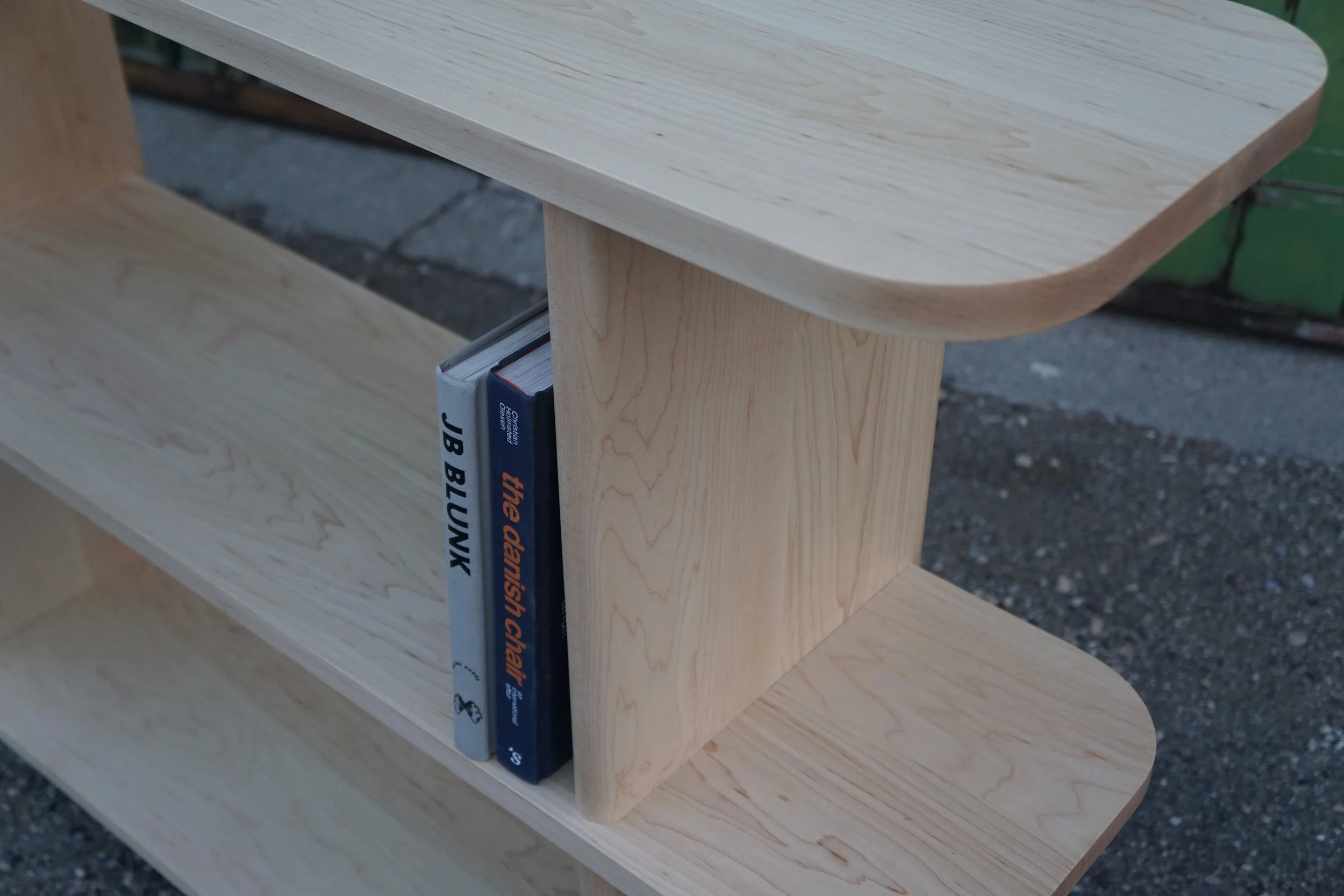 A wooden bookshelf with a curved top shelf, containing three books, outdoors on a gravel surface.